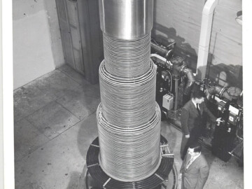 The 3.5 MV “home-made” vertical Van de Graaff accelerator that was installed in Bldg. 114 in the tower at the south end of the building. During operation the accelerator structure seen in the photo was contained in a thick-walled, bell jar-like, pressure vessel which has been hoisted up into the tower. The pressurized insulating gas allowed higher voltages to be achieved with a relatively short structure. Photo date unknown but the tower to house the accelerator was built in 1948. From left to right the scientists are E.B. Paul, B.W. Sargent and A.J. Ferguson. Photo source – NRC Archives. 102-1000