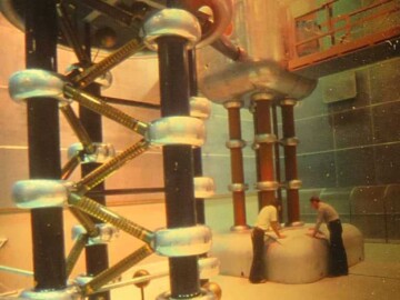 The injector of the HCTF. The Cockcroft-Walton 750 kV power supply is seen in the foreground while the high-voltage dome that housed the ion source and associated equipment is in the rear. Note that a drawbridge was used to allow access to the dome from ground elevation during maintenance times. This injector was operated until 1987 when it was dismantled and donated to the Canadian Museum of Science and Technology who hope to feature it in a future exhibit. L-R. D. Clements, H. Campbell.
<br /><br />
Photo source-AECL. 103-1000